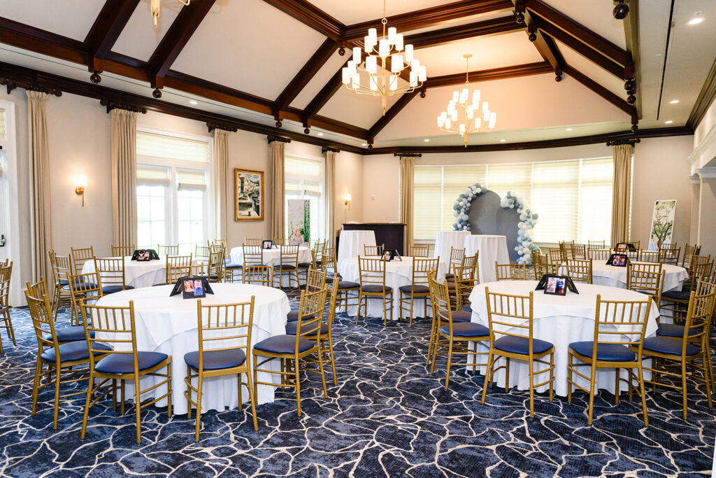 Ballroom set with round tables, gold Chiavari chairs, navy patterned carpet, and a blue-and-white balloon backdrop for Kaitlyn and Connor’s rehearsal dinner