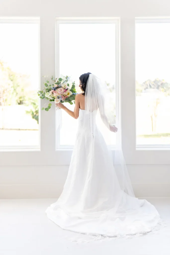 Bride in a lace gown holding her bouquet by bright windows before the ceremony
