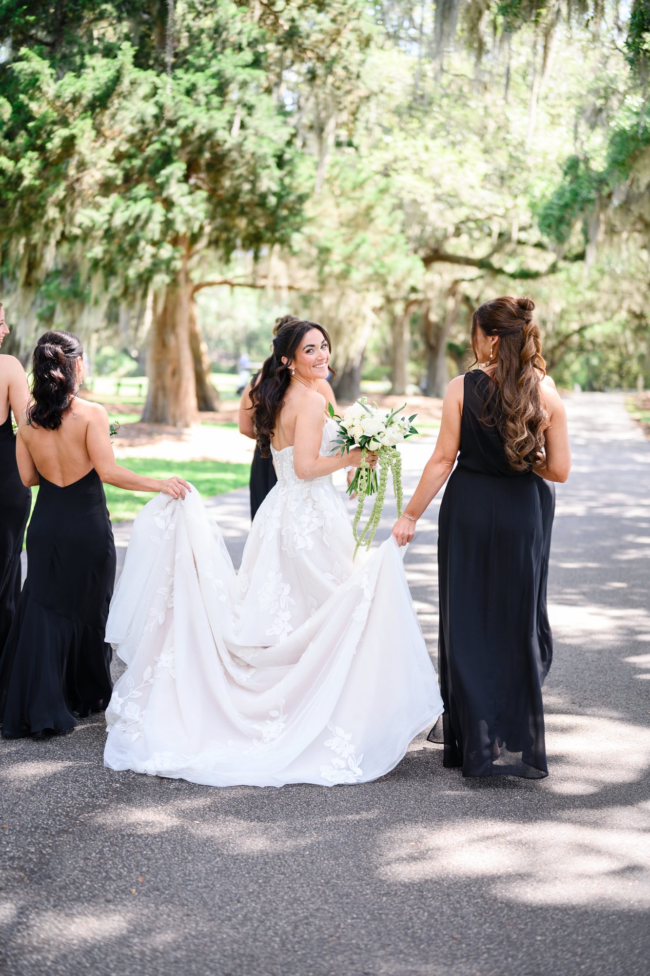 Bridesmaids in black gowns helping Ally with her train under the live oaks at Caledonia