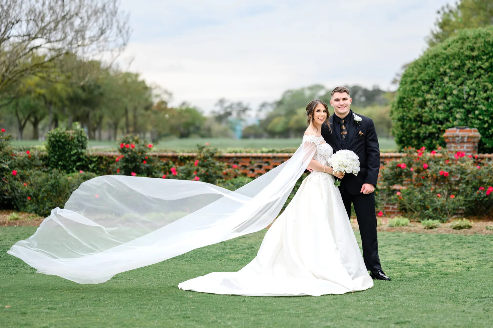 Kayla's cathedral veil catching the wind beside Austin on the Pine Lakes grounds