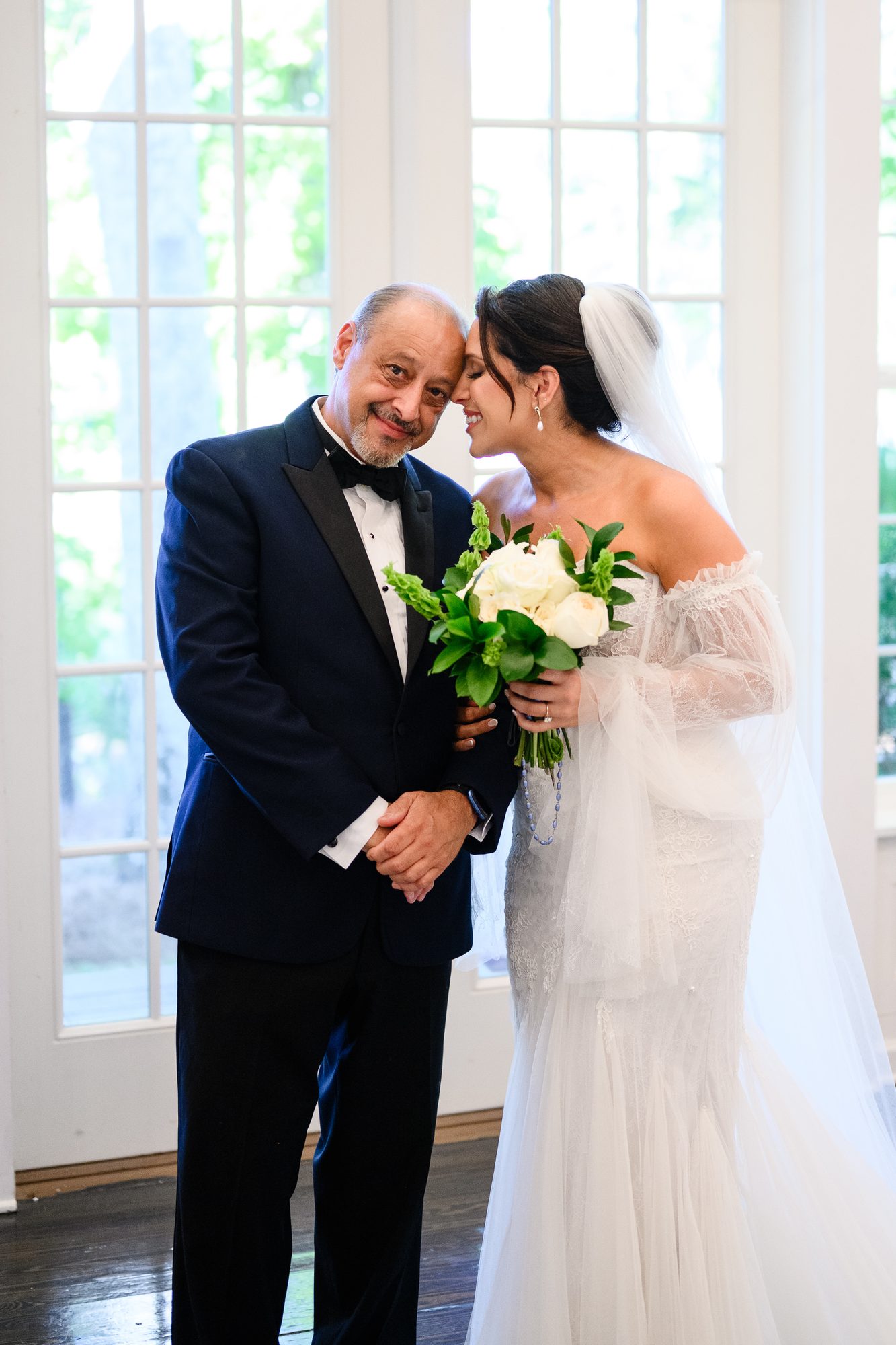 Shannon in a lace gown hugging her father by tall windows at The Village House